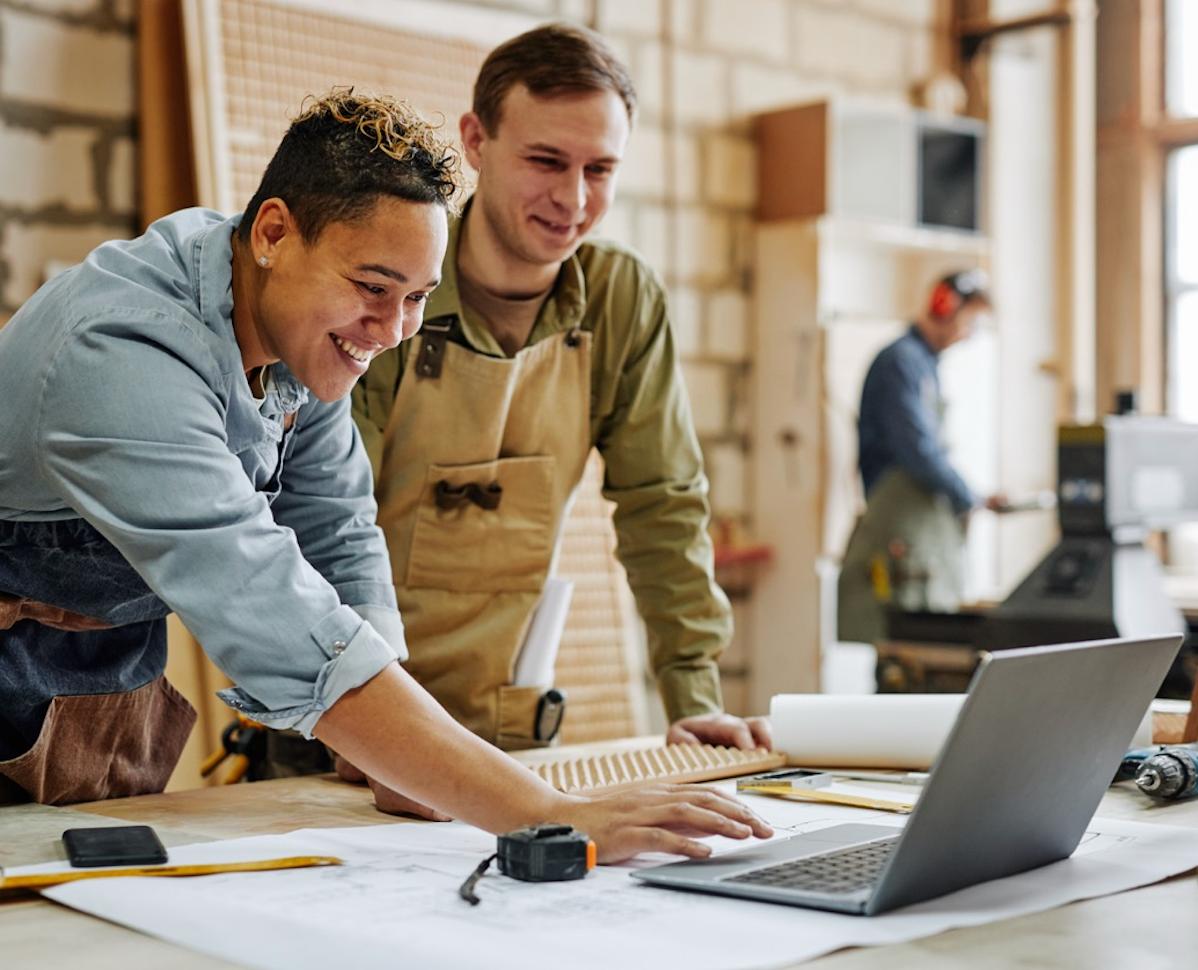 Two smiling woodworkers review designs on a laptop in a busy workshop.